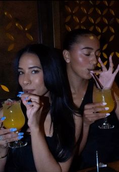 two women sitting at a table with drinks in front of their faces and one holding up her hand