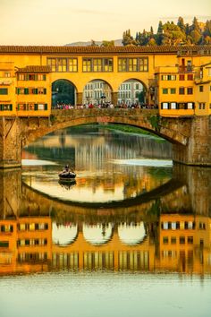 a small boat traveling down a river under a bridge with buildings on both sides and people walking across the bridge