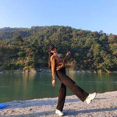 a woman in brown shirt and black pants walking on beach next to water with trees