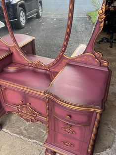 a pink vanity with an ornate mirror on the top and drawers below it, in front of a parked car