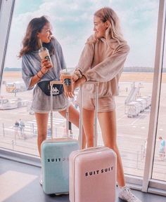 two women standing next to each other with suitcases in front of them and looking out the window