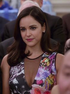 a woman in a floral dress sitting at a table with other people around her and looking into the camera