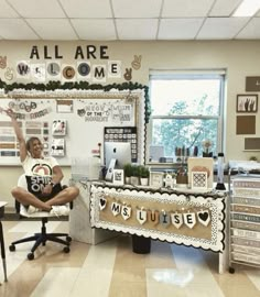 a woman sitting in a chair with her arms up and legs crossed, next to a bulletin board that says all are welcome