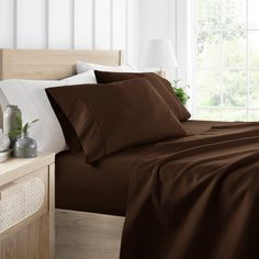 a bed with brown sheets and pillows in front of a large window next to a night stand