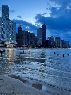 people are playing in the water at dusk on a beach with skyscrapers in the background