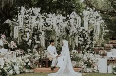 a bride and groom sitting on a bench in front of a flower covered wall with white flowers