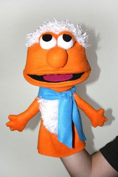 a hand holding an orange stuffed animal with white hair and blue scarf around it's neck