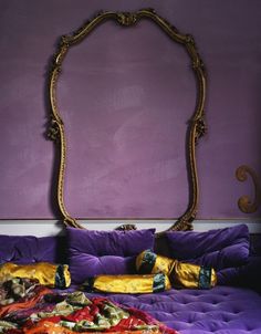 a bed with purple sheets and pillows in front of a gold framed mirror on the wall