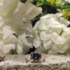 a blue diamond ring sitting on top of a stone wall next to white and green flowers