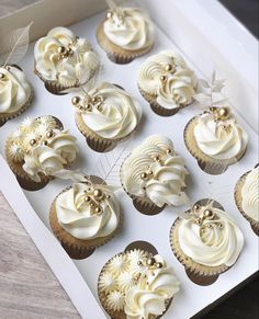 twelve cupcakes with white frosting and gold decorations in a box on a table