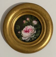 a painting of flowers in a gold frame on a white wall, with green leaves and pink roses