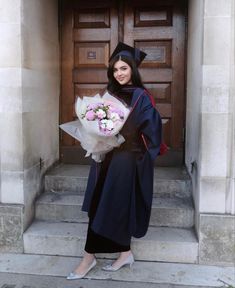 a woman standing in front of a door with flowers on her hand and wearing a graduation gown