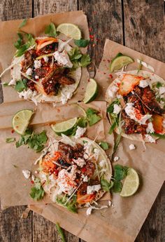 three tacos with meat, vegetables and cheese on brown paper wrappers next to lime wedges