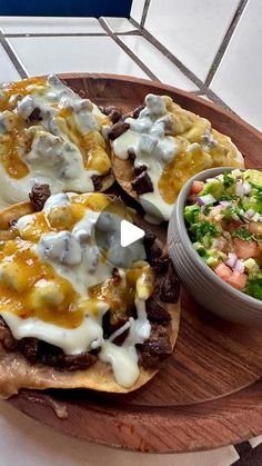 a wooden plate topped with three tacos covered in cheese and sauce next to a bowl of salad