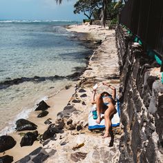 a woman laying on top of a blue and white towel next to the ocean in front of a stone wall