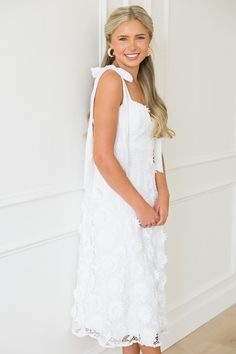 a woman standing in front of a wall wearing a white dress and smiling at the camera