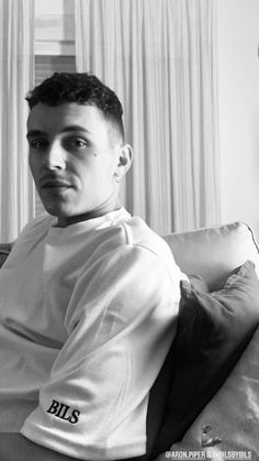 a young man sitting on top of a couch next to a window with curtains behind him