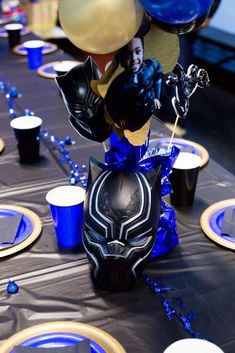 a table topped with blue and gold plates covered in black and white balloons next to batman masks