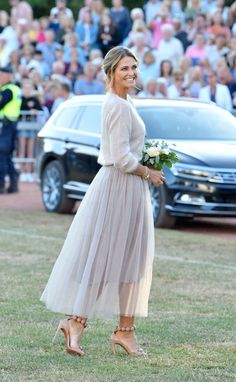 a woman in a dress is walking on the grass with flowers and people watching behind her