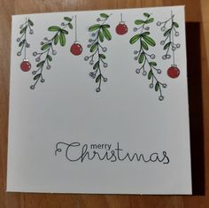 a merry christmas card with holly and berries on white paper, hanging from a wooden table