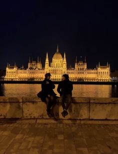 two people are sitting on a stone wall near the water at night, with a large building in the background