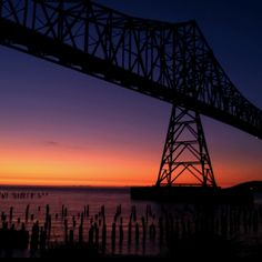 the sun is setting over the water under a large metal bridge that stretches into the distance