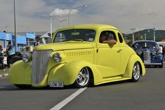 an old yellow car is driving down the street with other cars in the back ground