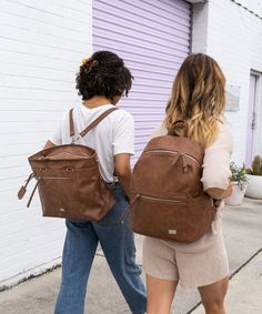 two women are walking down the sidewalk carrying backpacks on their backs and one has her back to the camera