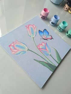 some watercolors are sitting on a table and there is a butterfly flying over the flowers