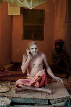 a man sitting on top of a bed talking on a cell phone with another man in the background