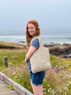 a woman carrying a crocheted bag walking down a wooden walkway next to the ocean