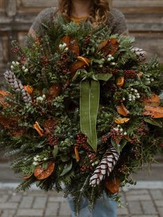 a woman holding a christmas wreath with pine cones, berries and greenery on it