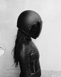 a woman wearing a black leather jacket with a motorcycle helmet on her head is standing in front of a wall