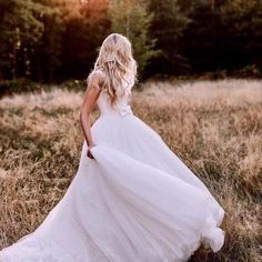 a woman in a wedding dress walking through the grass with trees in the back ground