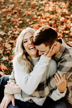 a man and woman cuddling in front of leaves on the ground with their arms around each other