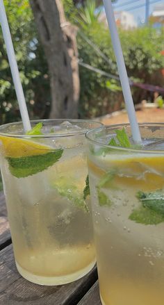 two glasses filled with lemonade and mint on top of a wooden picnic table outside