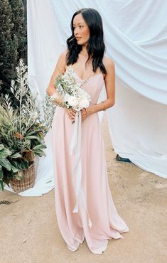 a woman standing in front of a white backdrop holding a bridal bouquet and wearing a pink dress