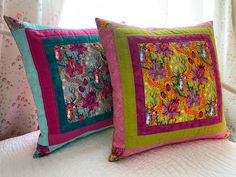 two colorful pillows sitting on top of a bed in front of a window with curtains