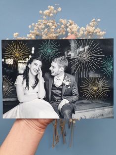 a hand holding up a photo with fireworks in the background and a bouquet of flowers