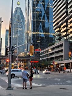 two people are standing on the street corner in front of tall buildings and traffic lights