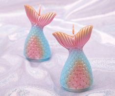 two little mermaid - like candles are sitting on a white sheet, one is pink and the other is blue