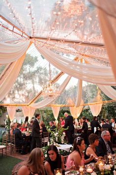 a group of people sitting at tables under a tent with white draping and chandeliers