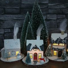 three small christmas houses are lit up in front of a stone wall and trees with snow on them