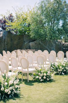 rows of white chairs with flowers on them in front of a rock wall and trees