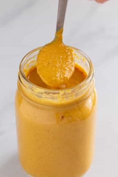 a person holding a spoon over a jar filled with orange colored liquid on a white surface