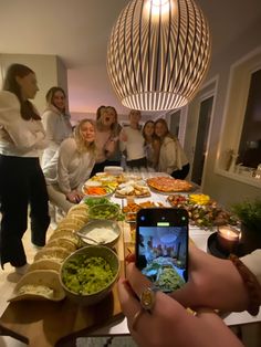 a group of people standing around a table with food on it and a cell phone in front of them