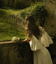 a woman in a white dress standing next to a stone wall with flowers on it