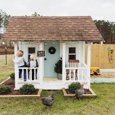 two children standing on the porch of a small white house with chickens in front of it