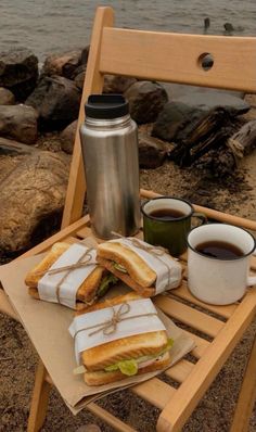 sandwiches and coffee sit on a wooden chair near the water with a cup of coffee in front of them