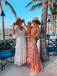 three women in dresses and hats standing next to each other on the beach with palm trees